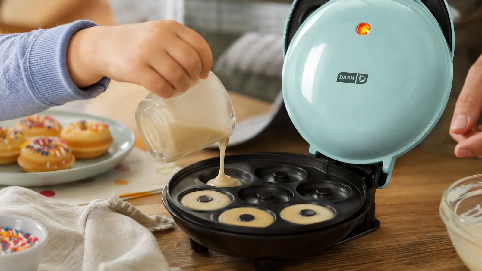 A child's hand pouring batter into a mini donut maker on a kitchen counter