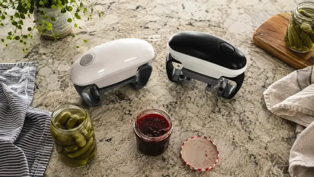 Two electric jar openers sitting side by side on a kitchen counter next to jars of pickles and jam