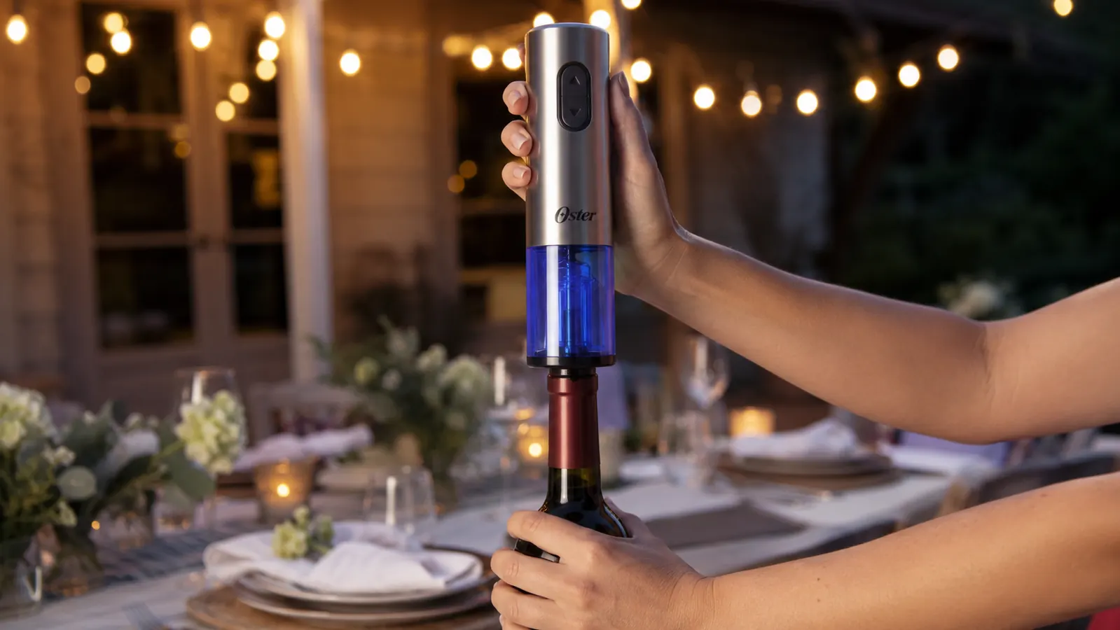 A hand placing the Oster cordless electric wine opener on top of an open wine bottle at an outdoor dinner table